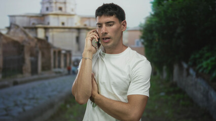 Young hispanic man holds smartphone to ear and cradles elbow on historic cobblestone street; concern.