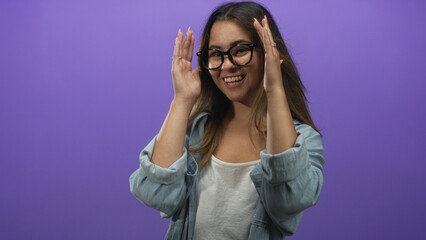 Young hispanic woman smiles and covers face with hands while adjusting glasses in studio purple set; playful curiosity.