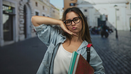 Young woman holding books and flashing a peace sign with her hand on a cobblestone street near a plaza and buildings; playful confidence.