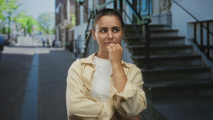 Woman with hand on chin standing on city street near stone steps and metal railings, wearing beige...