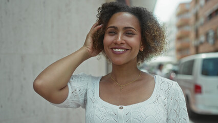 Woman smiling while touching hair on a city street, wearing white sweater and cross necklace on...