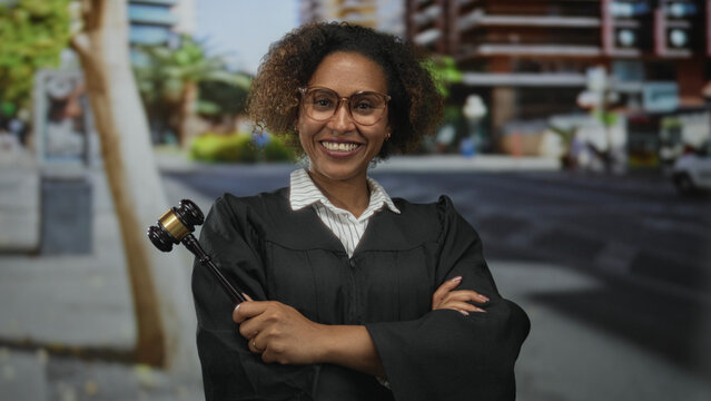 Woman judge holding gavel with crossed arms on street smiling, wearing robe and glasses; justice confidence.