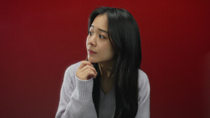 Woman thinking isolated against vivid red background wearing cozy sweater with thoughtful...