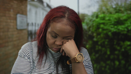 Woman rests cheek on hand wearing wooden watch in front of brick building beside hedge under...