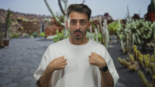 Man standing pointing finger to white shirt front in green park with cactus garden path; confidence.