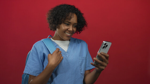 Young african american woman wearing scrubs holding smartphone and adjusting backpack strap in studio; happiness.