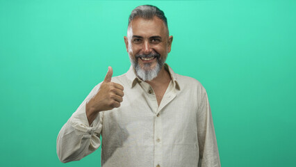 Middle aged man pointing finger at chest and giving thumbs up in green studio wearing beige linen shirt with a playful smirk; approval signal.