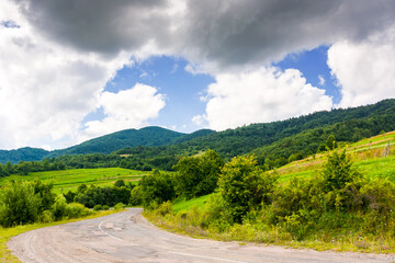 uzhanian pass in carpathian mountain landscape. old asphalt country road take turn downhill near forest. rural scene with field and haystacks in summer under cloudy sky. countryside travel background
