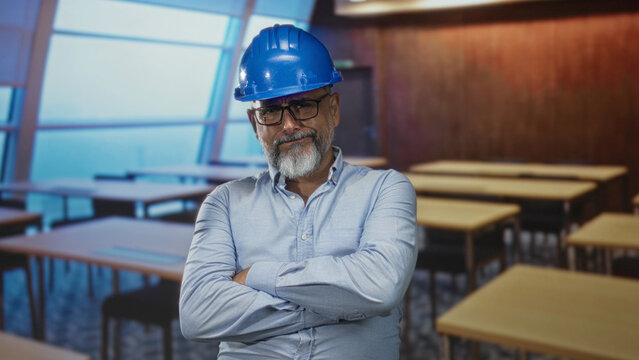 Man architect wearing blue hardhat with arms crossed in a building classroom; experience authority confidence.