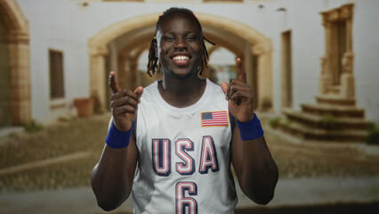 Man points index fingers upward on a cobbled street in an old town, smiling and wearing a usa...