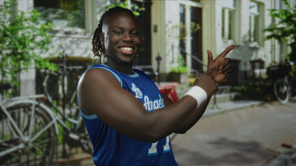 Man smiling points finger while showing bare arm on street in front of building and bicycles, wearing blue los angeles jersey; confident joy.