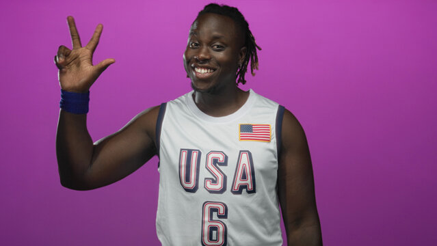 Man in usa basketball jersey shows thumbs up and raised hand in studio with magenta background;confidence celebration.
