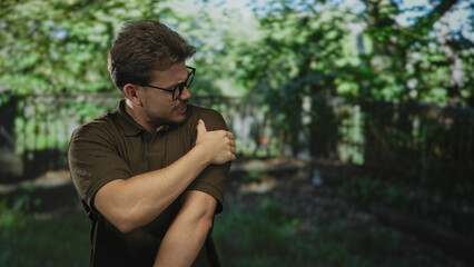 Man clutching bare shoulder and grimacing while wearing glasses and a brown polo shirt in a forest park setting; pain recovery.