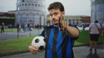Fototapeta premium Man holding soccer ball and pointing finger to camera in front of pisa tower building plaza; confidence ambition team spirit.
