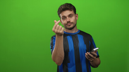 Man wearing blue striped jersey making finger heart gesture with smartphone in green studio; playful charm.