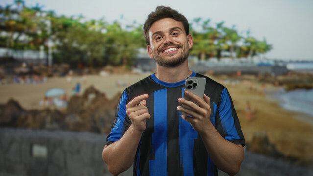 Young hispanic man points finger to smartphone on beach while smiling and holding phone in hands; happiness connection relaxation.