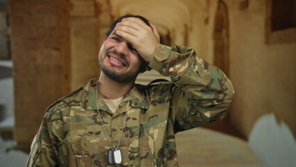 Man in camouflage uniform with visible dog tags and hand on forehead in building corridor, mouth...