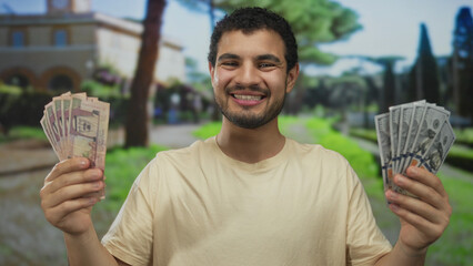 Smiling young hispanic man holds riyal and dollar bills with both hands in front of a building;...