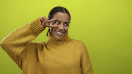 Young woman smiling and posing with a peace sign wearing a cozy sweater against a vibrant yellow...