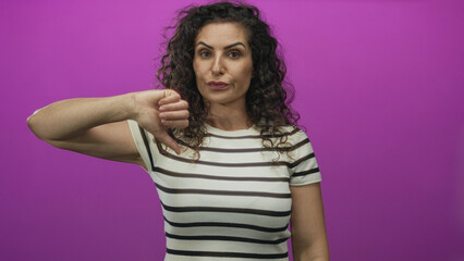 Hispanic woman giving a clear thumbs down gesture in purple studio setting; rejection disapproval dislike.