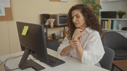 Obraz premium Young hispanic woman holds wrist while typing on black desktop keyboard at tidy office desk in modern building; work discomfort.