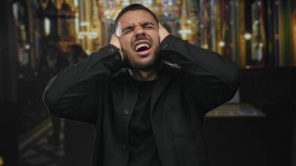 Young hispanic man in black shirt standing intensely covers ears with both hands in church building; anguish.