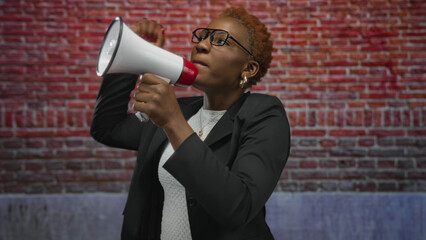 Young african american woman shouting into a megaphone against a red brick building while raising her fist and wearing glasses; empowerment advocacy solidarity activism.