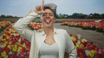 Woman makes l sign on forehead and sticks out tongue in studio with colorful tulip rows;...