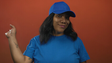African american woman in blue cap and t shirt waves hand in studio against orange wall; warm welcome.