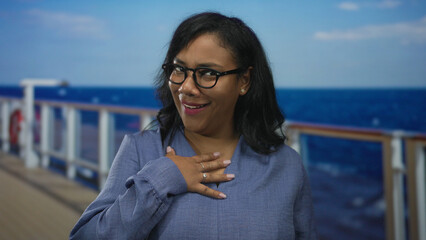 Woman wearing glasses holds hand on chest on cruise deck by ocean horizon with an awkward smile; surprise.