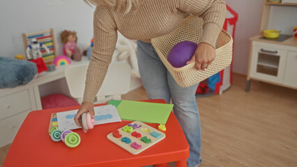 Woman arranging toys into a basket while holding purple ball in kindergarten building; nurturing...