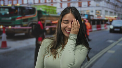 Fototapeta premium Young woman covering her eye with her hand on a city street in front of construction vehicles with traffic cones behind; shyness.