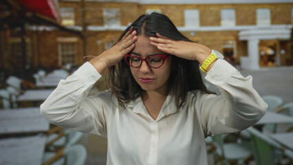 Fototapeta premium Woman wearing glasses in a restaurant terrace outside during daytime appearing thoughtful and pensive while dressed in a white shirt and holding her head outdoors.