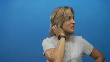 Young blonde woman in white shirt touches hair while raising arm in studio portrait; uncertainty...