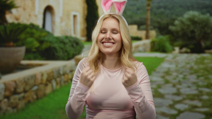 Young blonde woman with bunny ears smiling in a mediterranean stone house setting, exuding joy and playful energy amidst vibrant greenery, embodying a fun atmosphere.