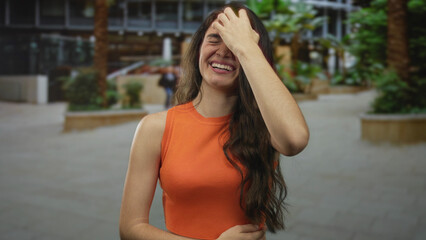 Woman in orange top laughing with hand in hair on street by building facade amid palm trees;...