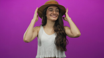 Young hispanic woman adjusting straw hat with closed eyes in purple studio; playful excitement.