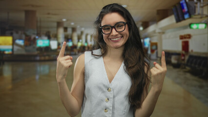 Woman wearing striped vest pointing both index fingers upward while smiling broadly in airport;...