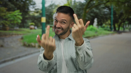Young hispanic man making rude gesture and smiling in a green park setting outdoors, expressing...