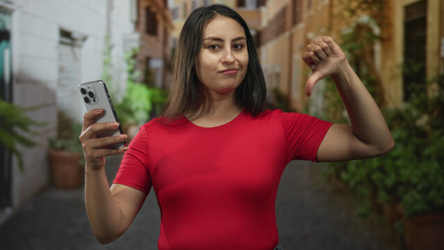Young hispanic woman holding smartphone with one hand and giving thumbs down with other, standing in narrow city street with potted plants; digital disapproval.