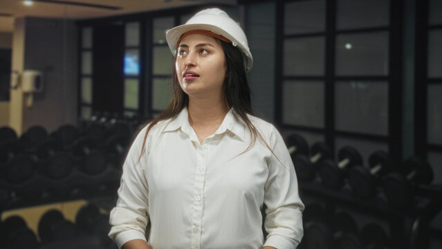 Woman architect wearing hardhat and white shirt looking up toward ceiling and gym dumbbells in building; confidence planning.