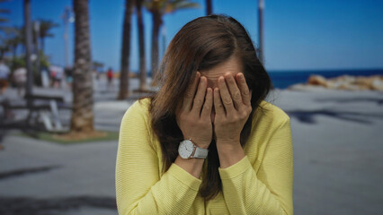 Fototapeta premium Woman covering face in anxiety while standing on sunny seaside promenade with palm trees.