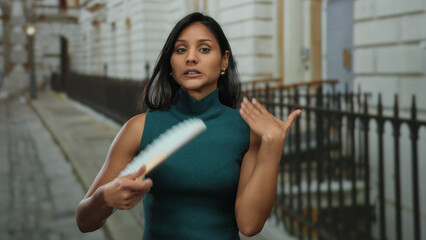 Latin woman with black hair fanning herself on a street, wearing a teal sleeveless top, expressing coolness on a breezy urban afternoon, showcasing natural beauty in the city.