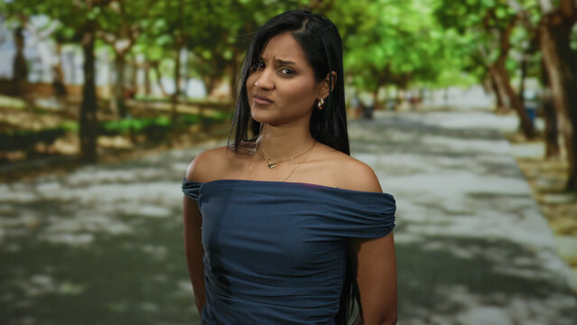 Woman with skeptical expression stands outdoors in vibrant park wearing blue dress surrounded by lush green trees showing a thoughtful demeanor in a natural setting