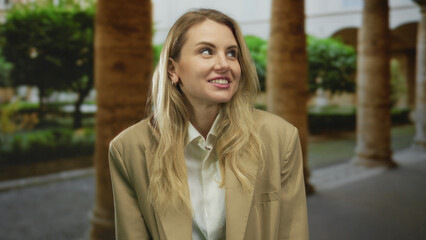 Fototapeta premium Woman smiling in outdoor setting with blonde hair and beige jacket, surrounded by lush green plants and architectural columns on a bright day.