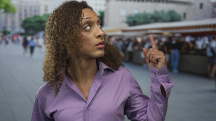 Urban woman in purple shirt pointing finger with raised eyebrow on bustling street beside souvenir stalls; direction attention assertion confidence.