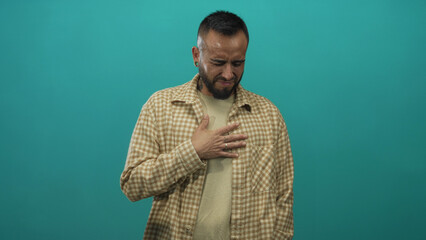 Young man bearded in checkered shirt clutching chest with a pained grimace in teal studio; pain...