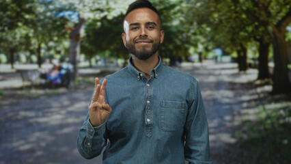 Man in denim shirt holds out open palm and points finger while smiling on a tree lined street with benches and dappled sunlight; contentment.