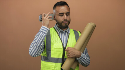 Man engineer wearing high visibility vest holding rolled blueprint tubes and smartphone to ear in peach studio; planning focus.