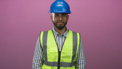 Man engineer wearing blue hardhat and neon safety vest smiles and taps wrist in pink studio; safety awareness confident readiness.
